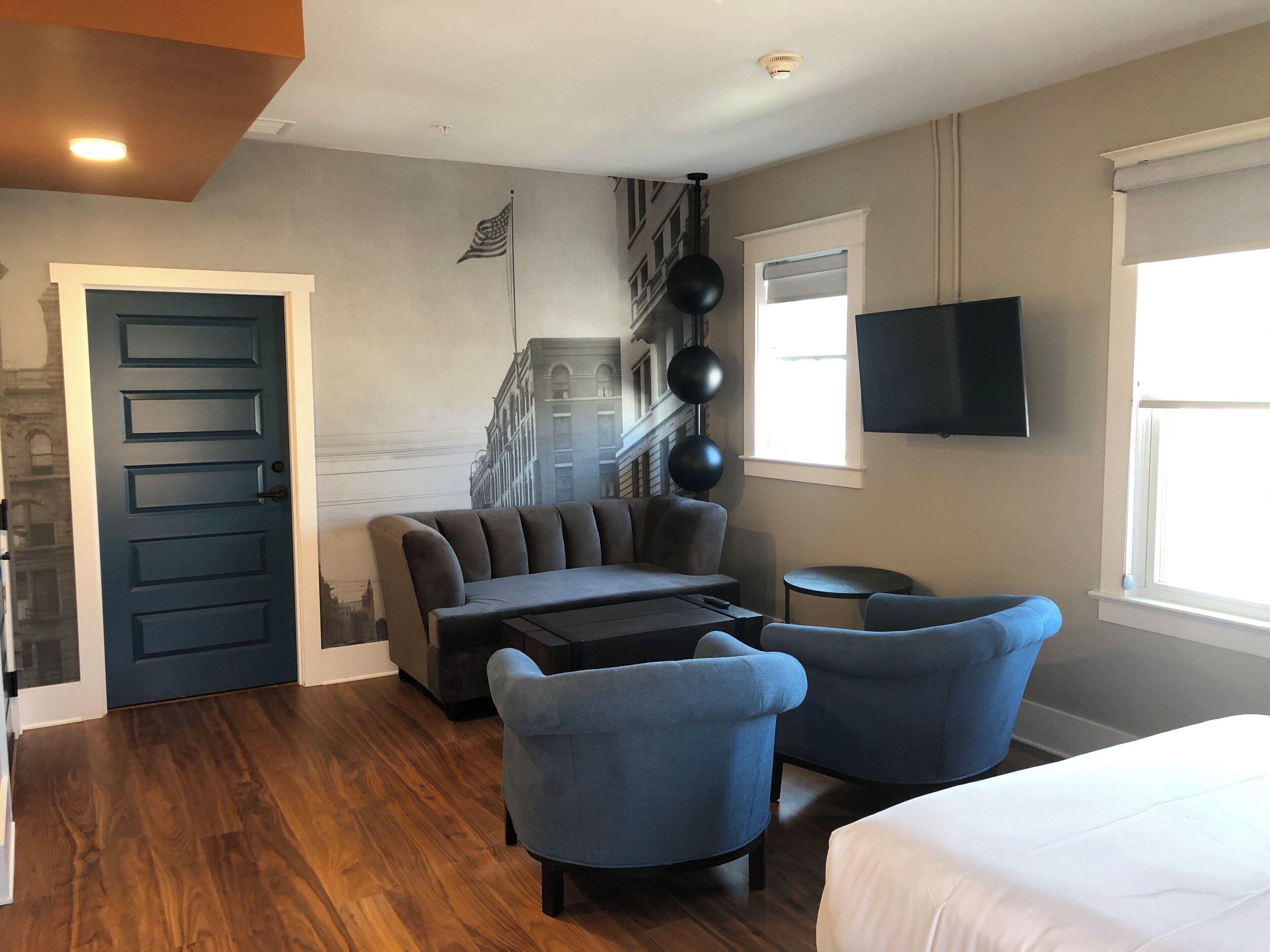 A hotel room with a sofa, two armchairs, a small table, a bed, a TV, a window, and a door.