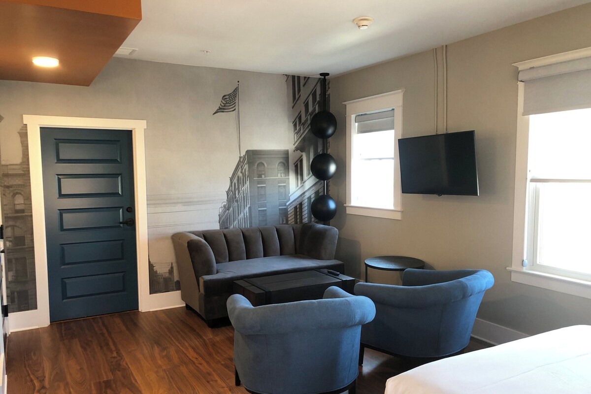 A hotel room with a sofa, two armchairs, a small table, a bed, a TV, a window, and a door.