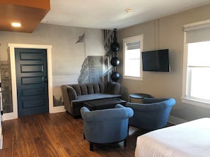 Une chambre d’hôtel avec un canapé, deux fauteuils, une petite table, un lit, une télévision, une fenêtre et une porte.