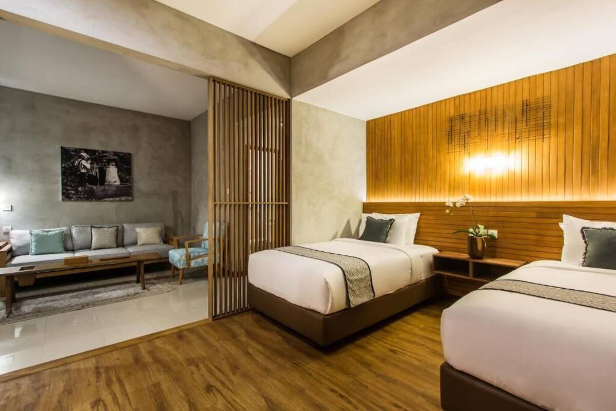 A modern hotel room with two beds, a wooden headboard, a large window, and a grey sofa.