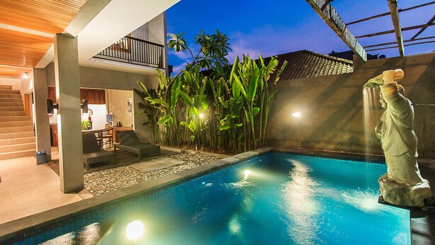A swimming pool with a statue, surrounded by plants and a modern building with a balcony. - Kuta Legian Villa (Kuta)