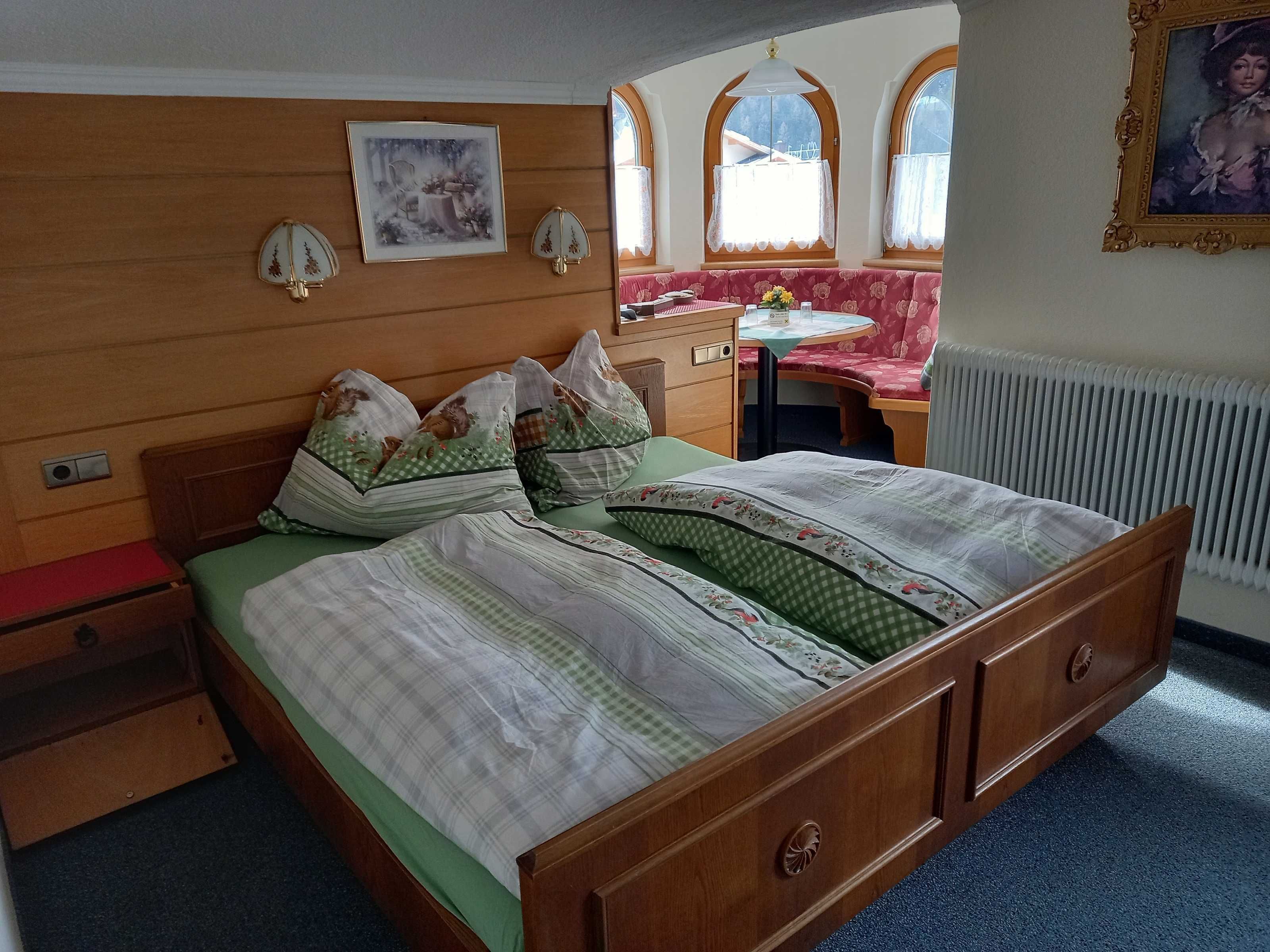 Ein hölzernes Bett mit grüner Bettwäsche, ein kleiner Nachttisch, ein Sessel, eine Heizung und ein Fenster mit Blick.