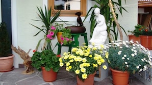Un’aiuola con piante in vaso davanti a un edificio con una finestra, una statua e uno sgabello verde.