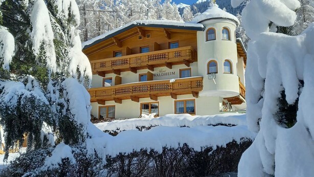 Um edifício de vários andares com uma placa indicando 'Haus Waldheim', cercado por árvores cobertas de neve e um céu azul límpido.