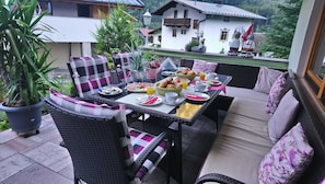 Een terras met rieten meubilair, een tafel gedekt met borden, brood en drankjes, omgeven door potplanten en met uitzicht op een bergdorp.