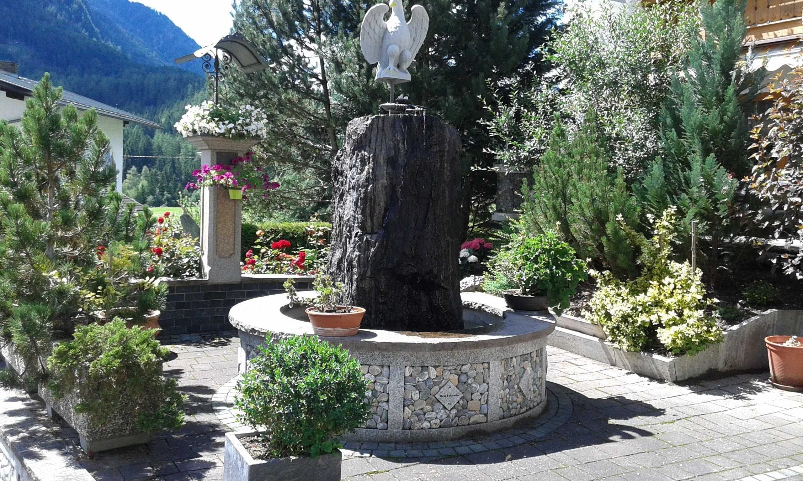 Ein Brunnen mit einer Vogelstatue, umgeben von Topfpflanzen und einer Steinmauer.