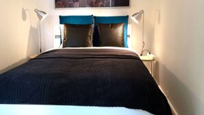 A neatly made bed with a dark bedspread and two pillows, a cityscape view, and a nightstand with a lamp. - StayPlus Central Apt Balcony (Oslo)