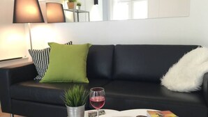A modern living room with a black leather sofa, a white coffee table, a magazine, a glass of red wine, and a small potted plant. - StayPlus Central Apt Balcony (Oslo)