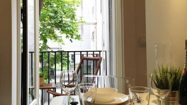 A dining area with a table set for two, a view of a balcony with a railing, and a potted plant on the table. - StayPlus Central Apt Balcony (Oslo)