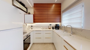 Eine moderne Küche mit weißen Schränken, einer Holzwand, einem Einbauofen, einer Kaffeemaschine und einem Fenster mit Jalousien.