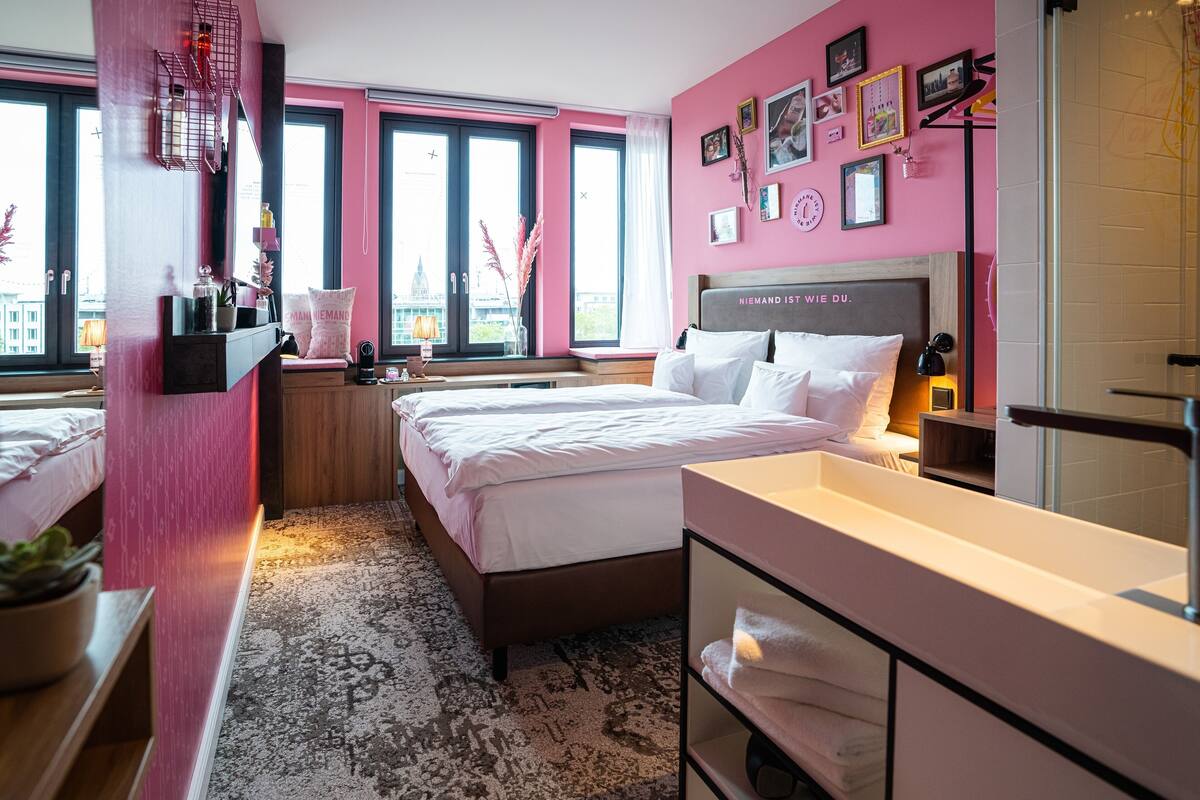 A modern hotel room with a large bed, a vanity with a mirror, and a view of the city through large windows.