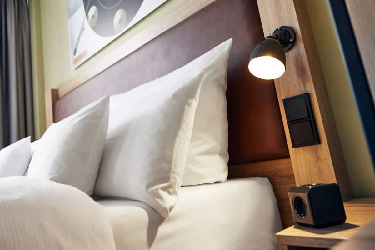 A neatly made bed with white linens and a wooden headboard. There is a wall-mounted light fixture on the headboard and a small speaker on the nightstand beside the bed.