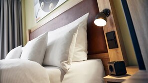 Una cama bien tendida con sábanas blancas y un cabecero de madera. Hay una lámpara de pared en el cabecero y un altavoz pequeño en la mesita de noche junto a la cama.