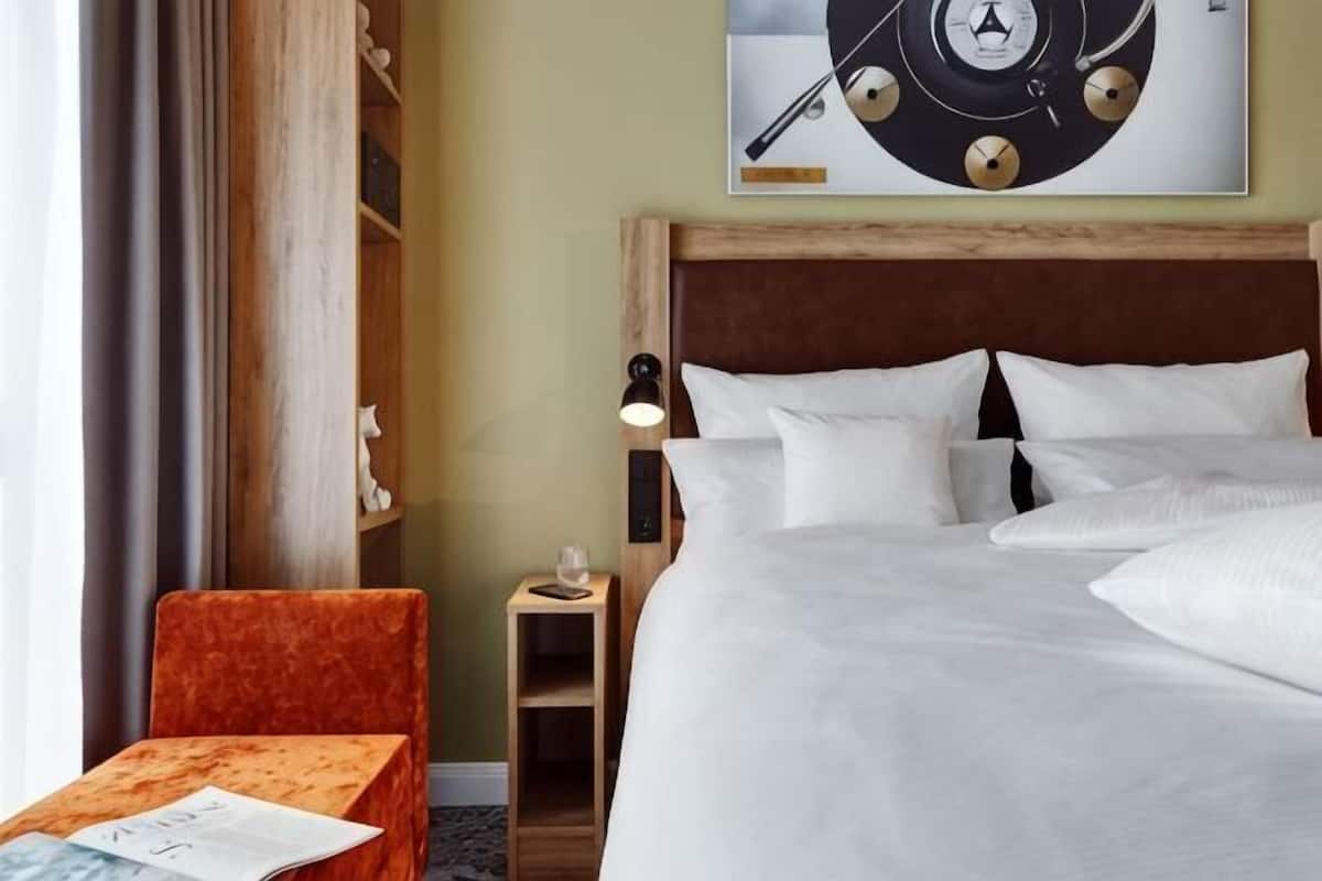 A hotel room with a bed, a nightstand, a chair, a clock, and a window with curtains.