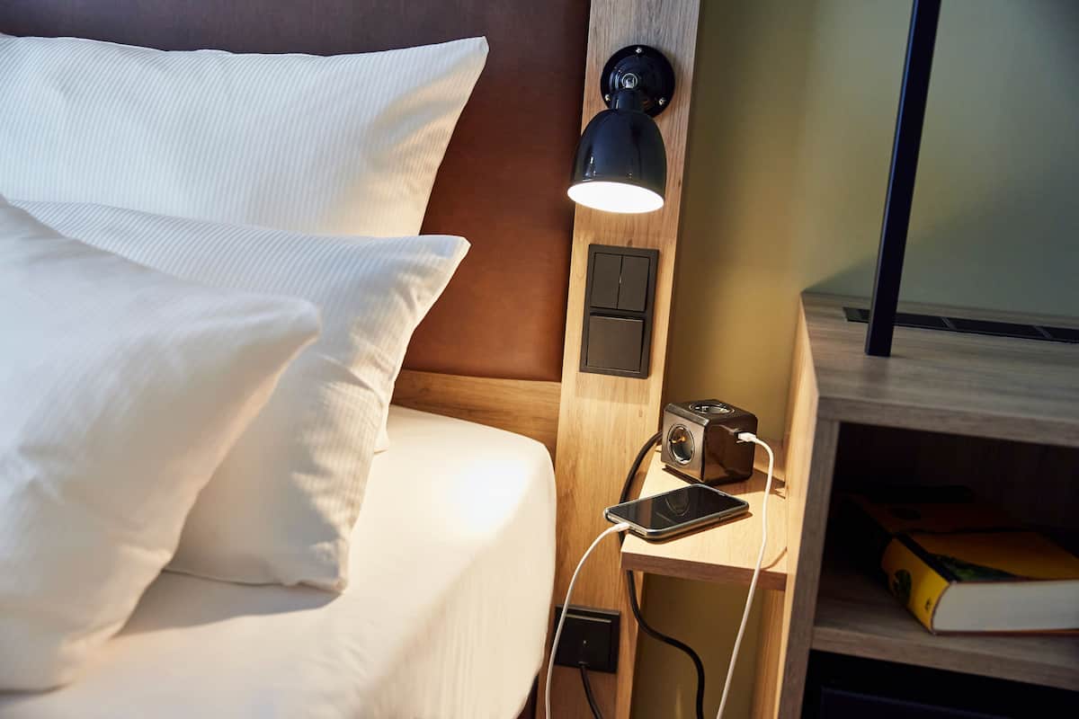 A neatly made bed with white linens, a bedside lamp, a smartphone, and a small wooden nightstand with a book and a camera.