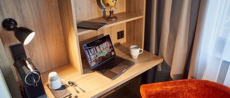 Un escritorio de madera con una computadora portátil, una cafetera y llaves, un globo terráqueo, y una silla roja en una habitación con paredes de madera y una ventana con cortinas.
