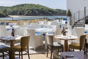 Un área de comedor al aire libre con mesas y sillas, con vistas a una playa y colinas.