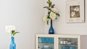 A dining area with a white table, two blue vases with white flowers, a cabinet with blue glassware, and a framed artwork on the wall. - Apartamento&piscina cerca de Barcelona (Ripollet)