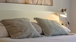 A neatly made bed with striped pillows and a white headboard. A wall-mounted lamp provides light, and a power outlet is visible. - Apartamento&piscina cerca de Barcelona (Ripollet)