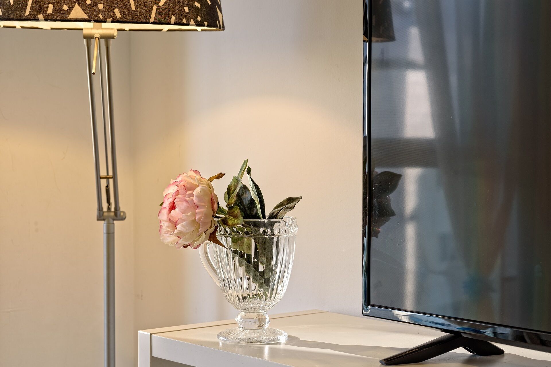 A clear glass vase with a pink flower and green leaves on a white surface next to a flat-screen TV.
