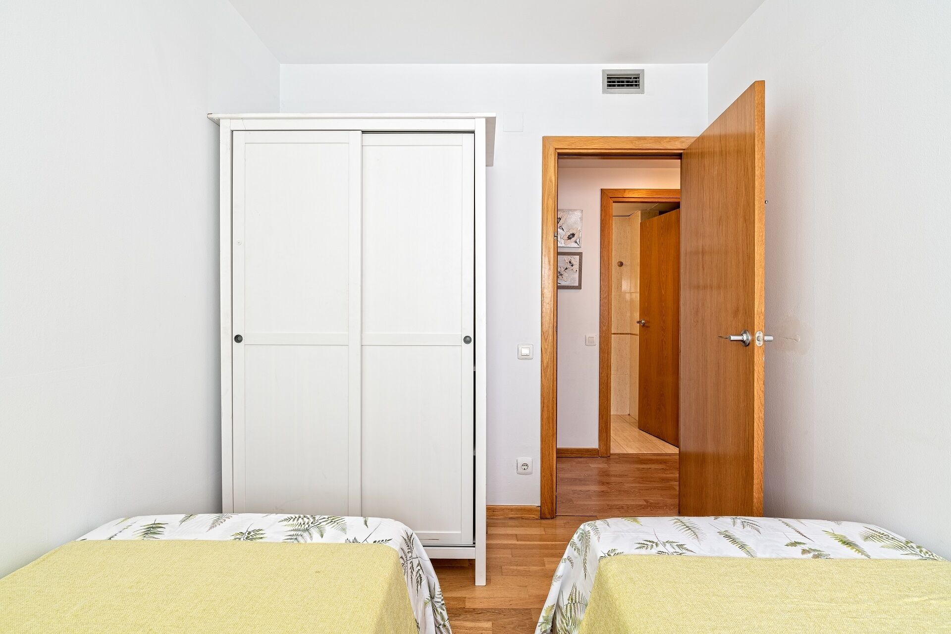 Una habitación pequeña y limpia con dos camas individuales, un armario blanco y una puerta de madera que da a otra habitación.