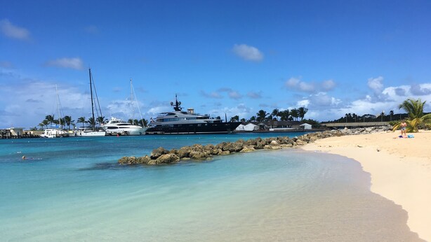 A beach with clear turquoise water, a sandy shore, and yachts docked nearby.