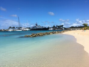 A beach with clear turquoise water, a sandy shore, and yachts docked at a marina.