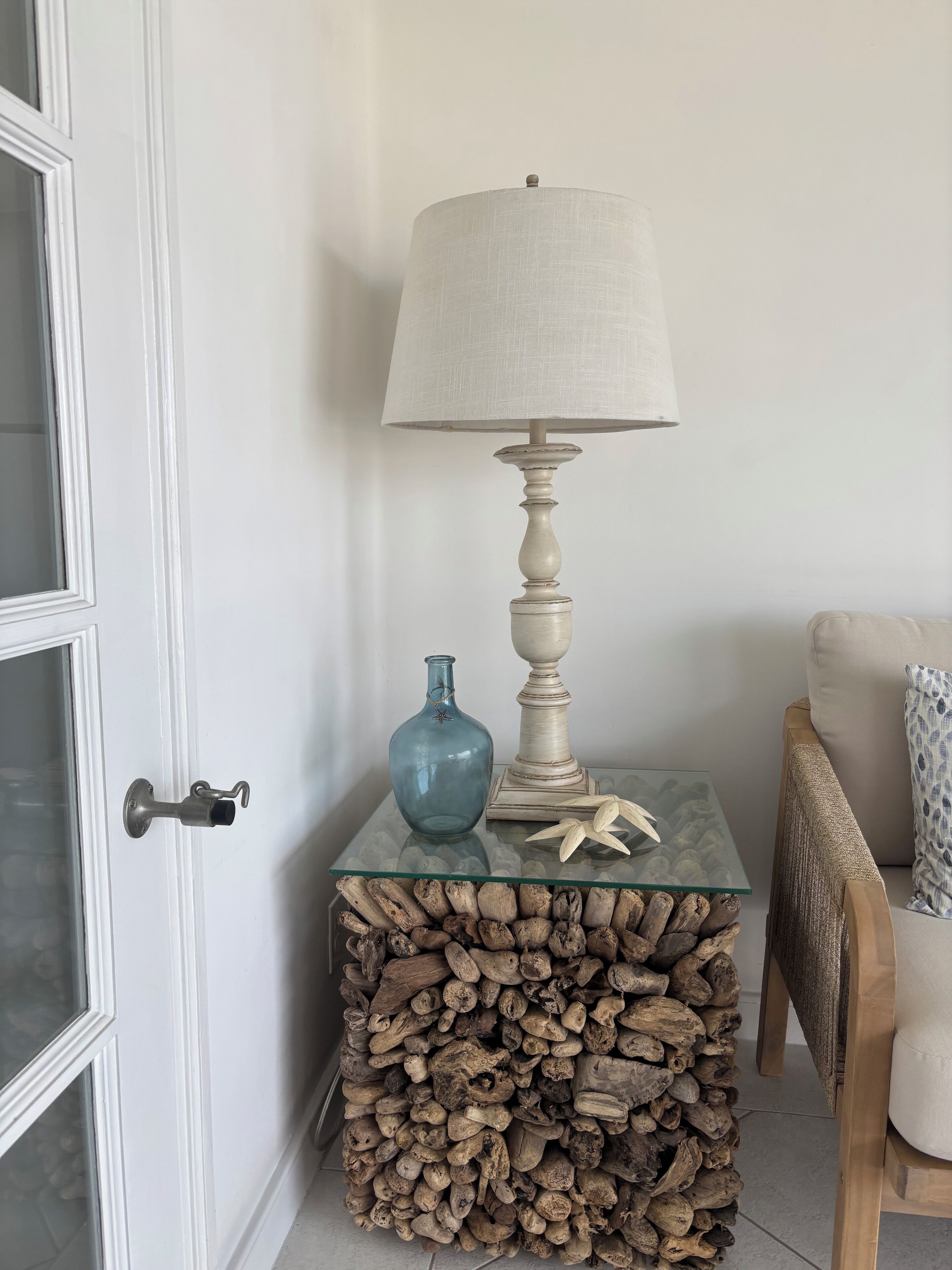 A corner with a white door, a glass table with a lamp, a blue vase, and a stack of logs.