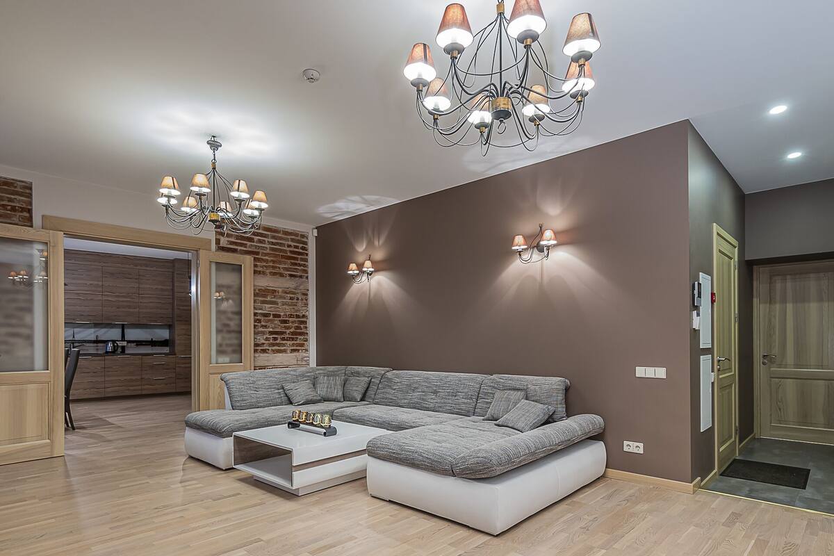 A modern living room with a sectional sofa, a coffee table, a chandelier, and a brick wall.