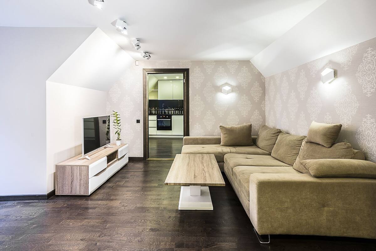 A modern living room with a sofa, a coffee table, and a TV on a wooden stand. There is a plant on the stand and a patterned wallpaper on the walls.