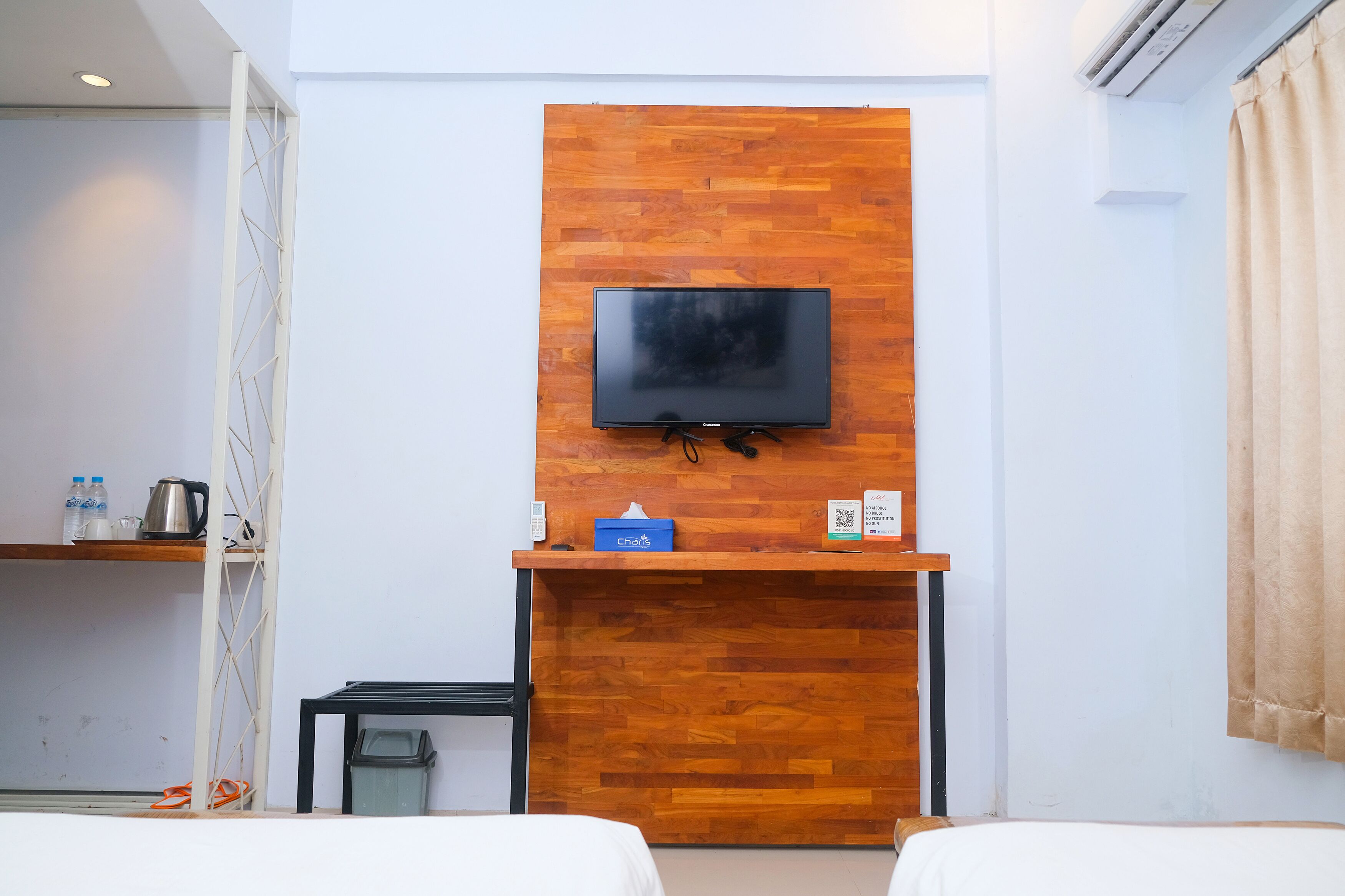 A hotel room with a wooden accent wall, a flat-screen TV mounted above a wooden desk, a bed, and a small trash bin.