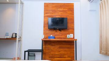 A hotel room with a wooden accent wall, a flat-screen TV mounted above a wooden desk, a bed, and a small trash bin.