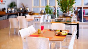 A dining area with white wicker chairs, a wooden table with a meal and drinks, and large windows in the background.