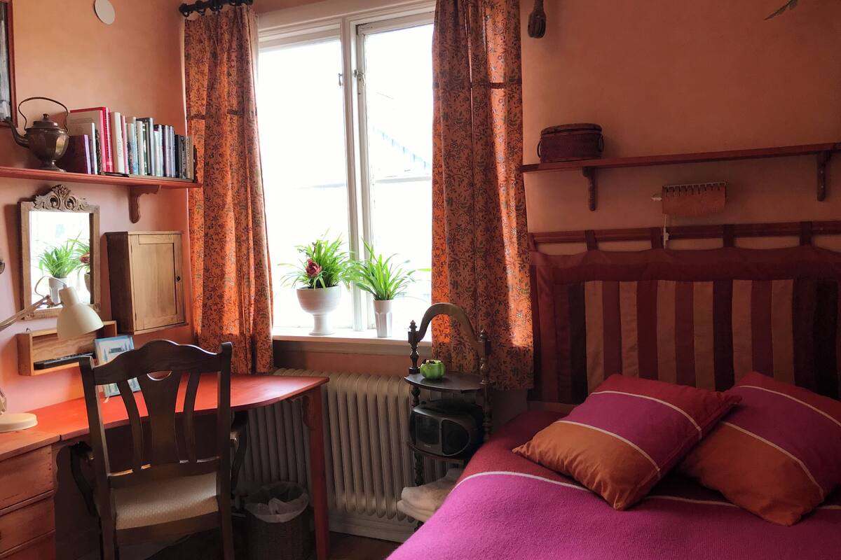 Un dormitorio acogedor con cama de madera, un escritorio y una silla. Se ve una ventana con cortinas, un radiador y un estante con libros y un hervidor.