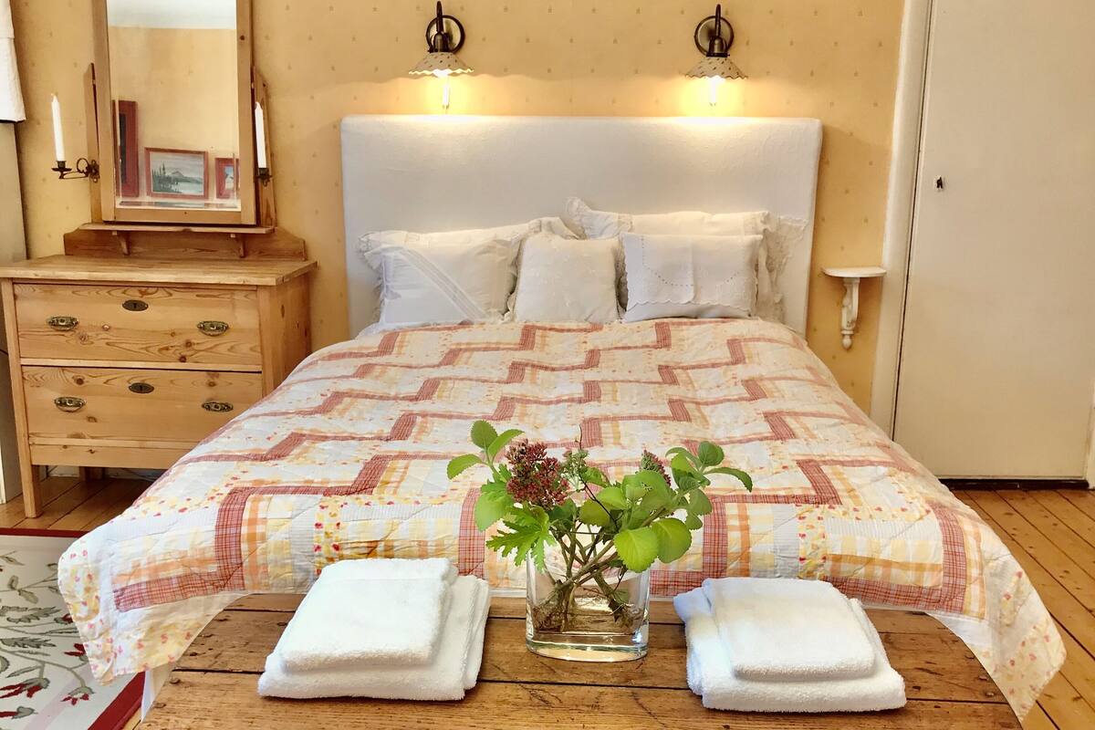 Un dormitorio con un cofre de madera, una cama con colcha, dos lámparas de pared, una mesita de noche con espejo y una pequeña mesa con una planta.