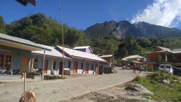 A small roadside building with a dirt ground, surrounded by greenery and mountains.