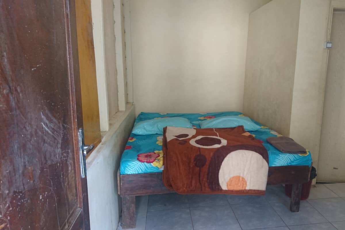 A small, narrow room with a bed against the wall, a wooden door, and a tiled floor.