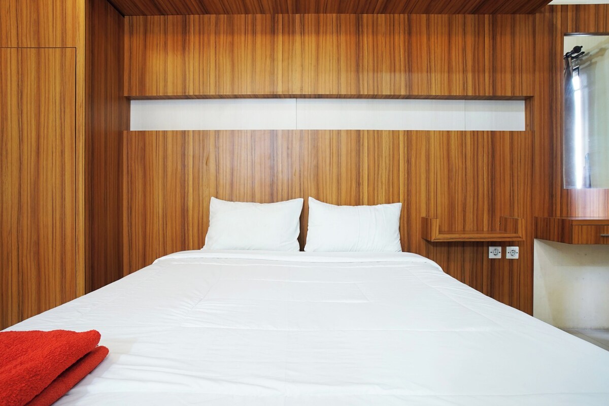 A neatly made bed with white linens and a red blanket, set against a wooden wall with a built-in shelf above it.