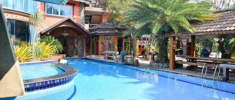 Una piscina con borde de azulejos, rodeada por un edificio con balcón y una zona cubierta con asientos.