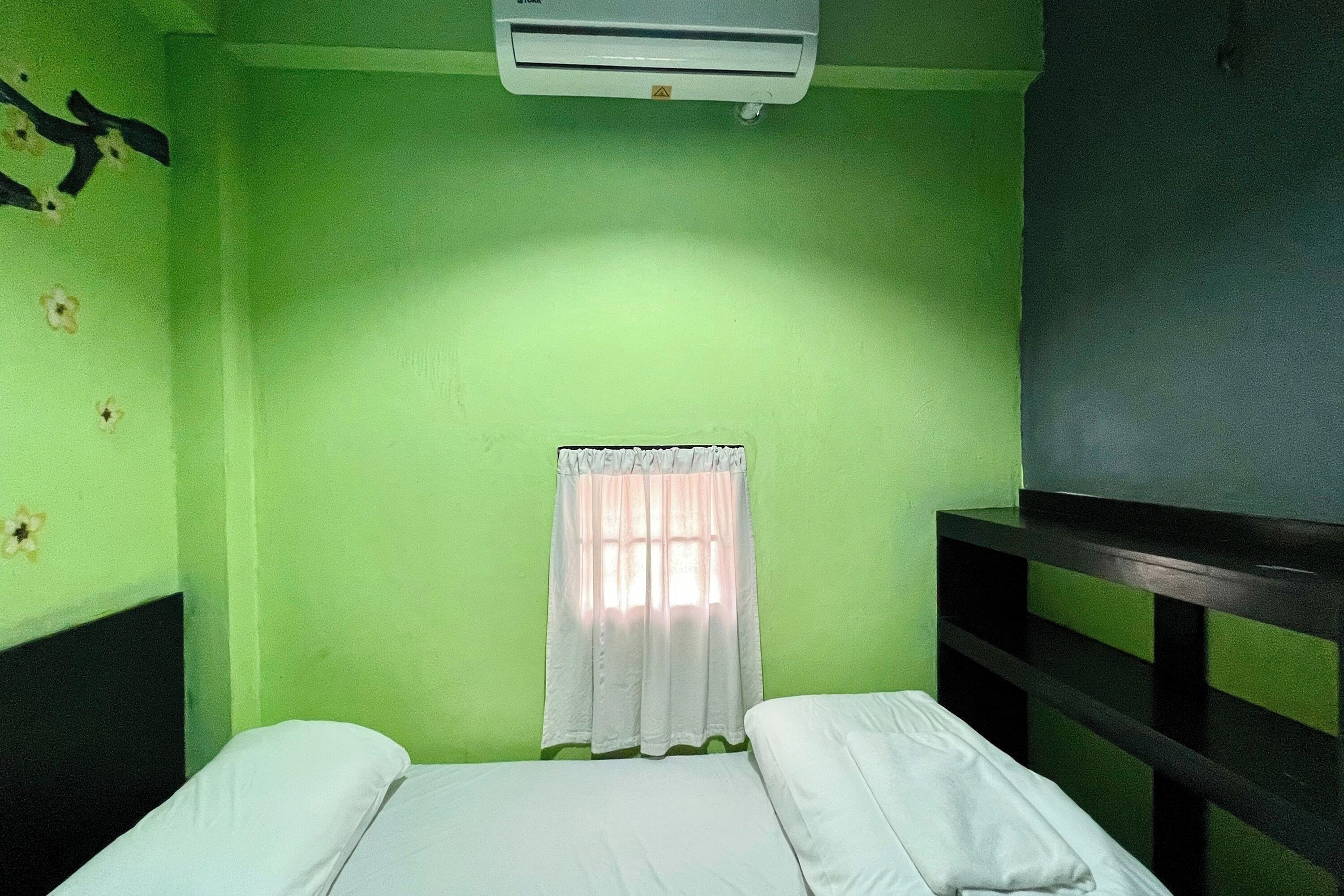 A room with a bed, a window with a white curtain, and an air conditioning unit on the wall.