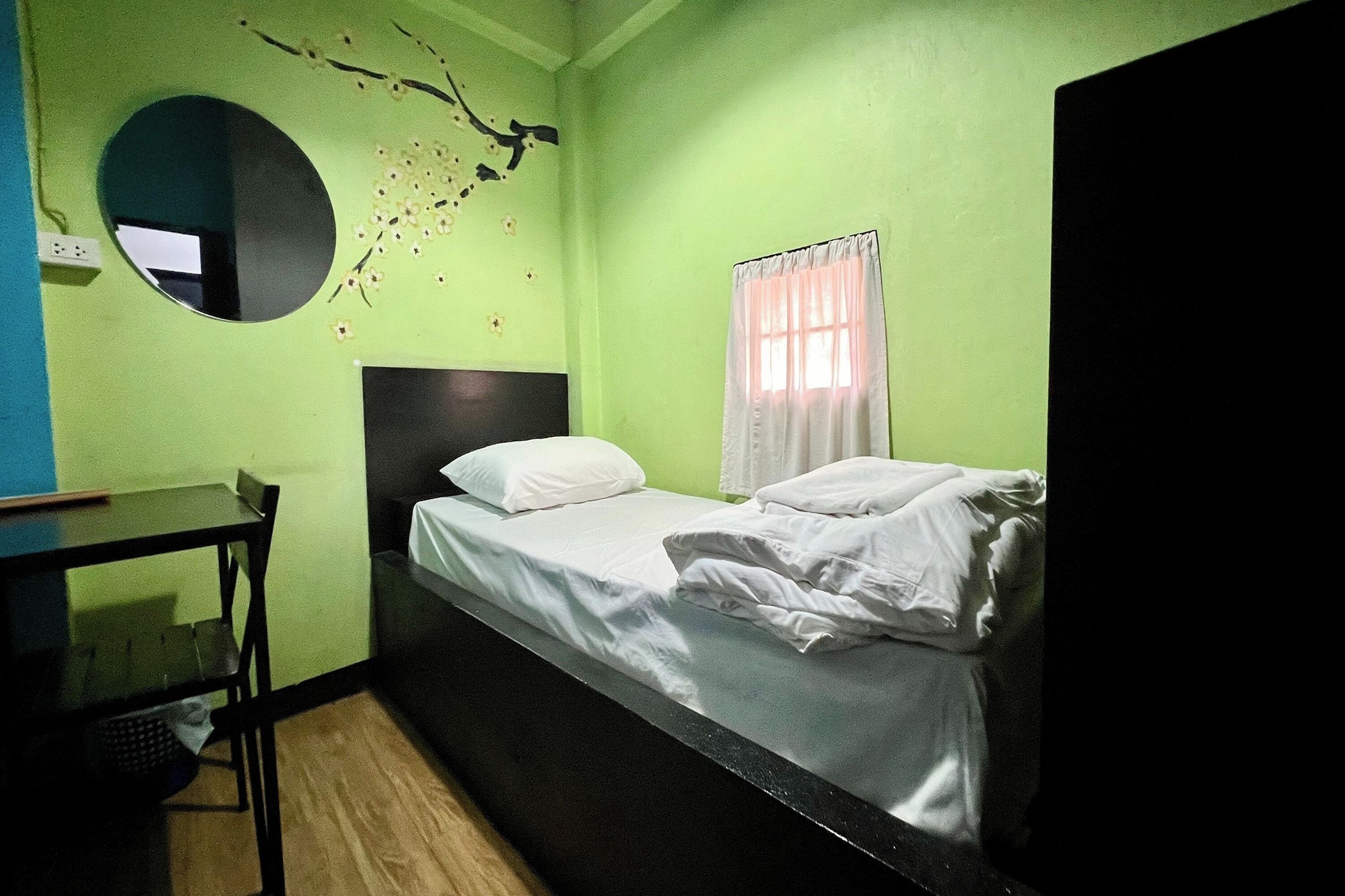 A small, single-bed room with a round mirror, a bedside table, and a window with curtains.