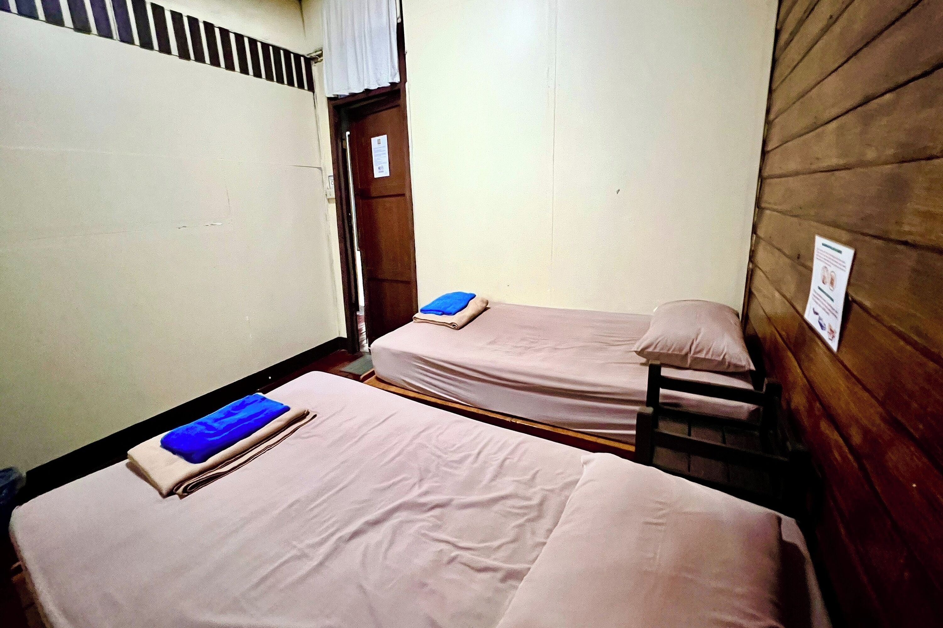 A small, compact room with two single beds, each with a pillow and a folded blanket. There is a wooden door and a wall-mounted sign.