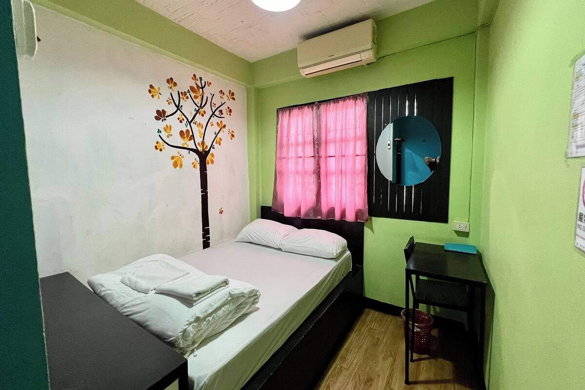 A small, single-bed room with a bed, a nightstand, a mirror, and a window with pink curtains.