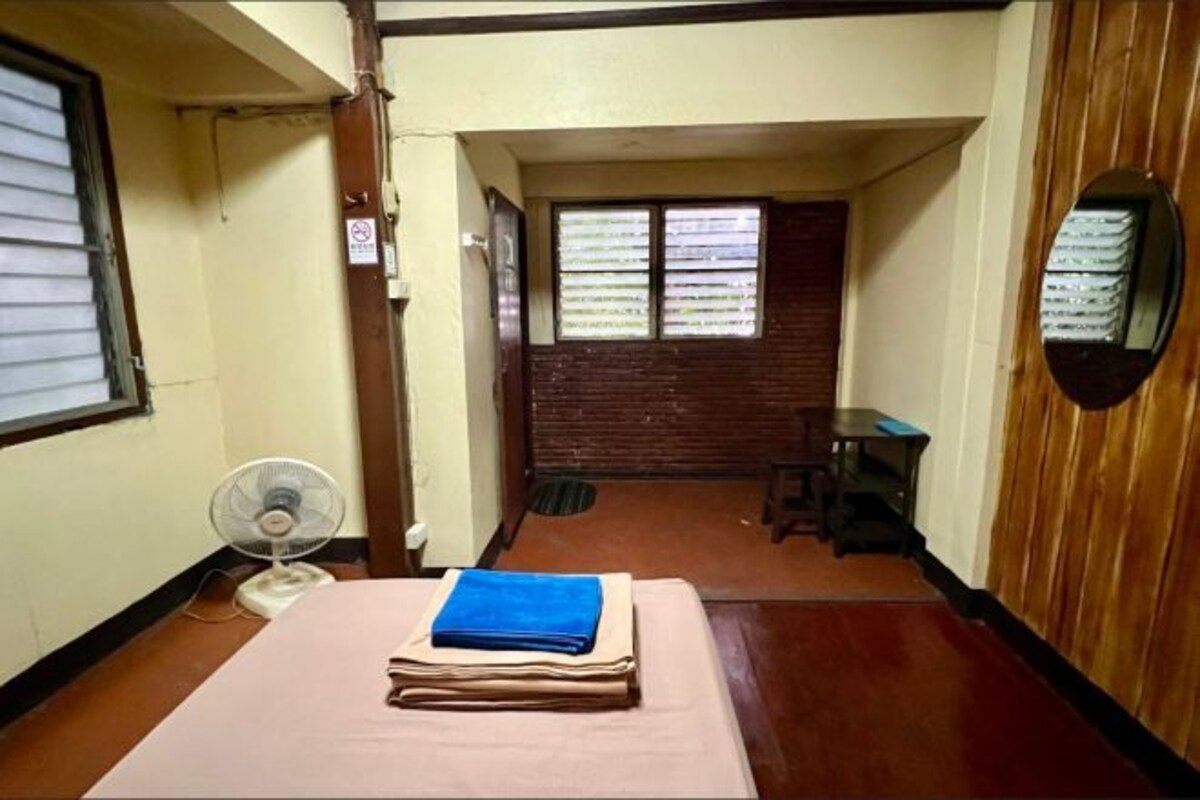 A narrow room with a bed, a fan, a small table, and a window with blinds.