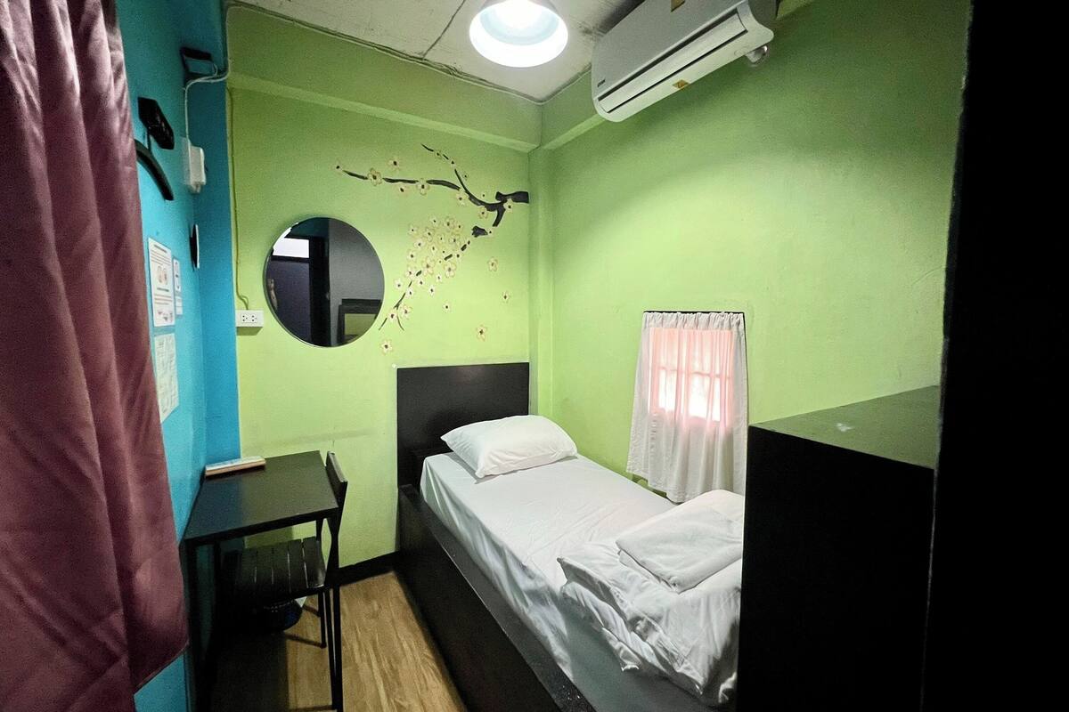 A small, single-bed room with a round mirror, a small table, and a window with curtains.