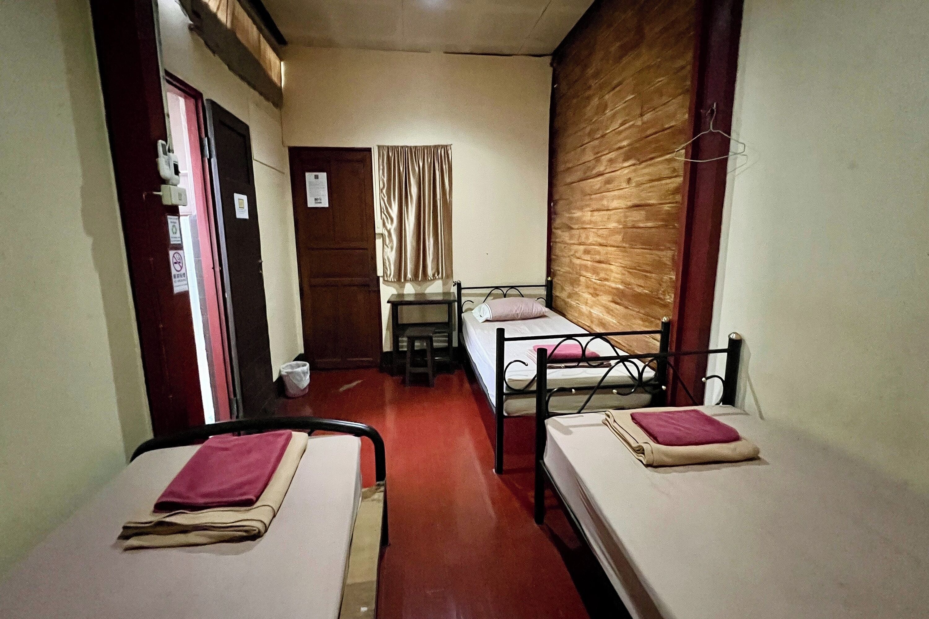 A narrow hotel room with two single beds, a small table, a chair, and a window with curtains.