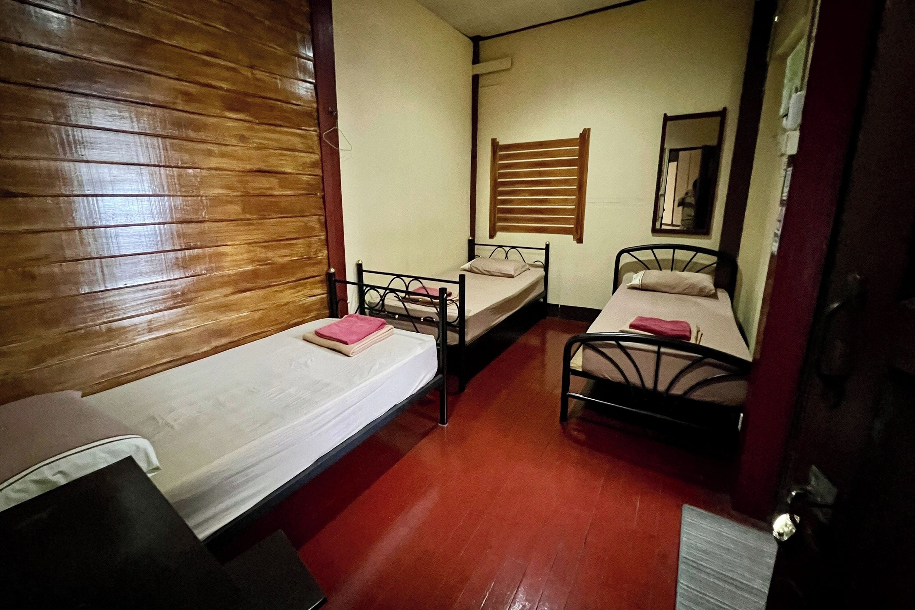 A small, compact room with two single beds, a wooden headboard, a window with blinds, and a mirror on the wall.