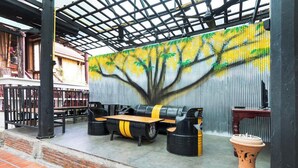 A covered outdoor seating area with a mural of a tree on the wall, a wooden table, and two black stools with yellow accents. - Baan Lanna At Phrasingh (Chiang Mai)