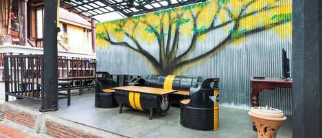 A covered outdoor seating area with a mural of a tree on the wall, a wooden table, and two black stools with yellow accents.