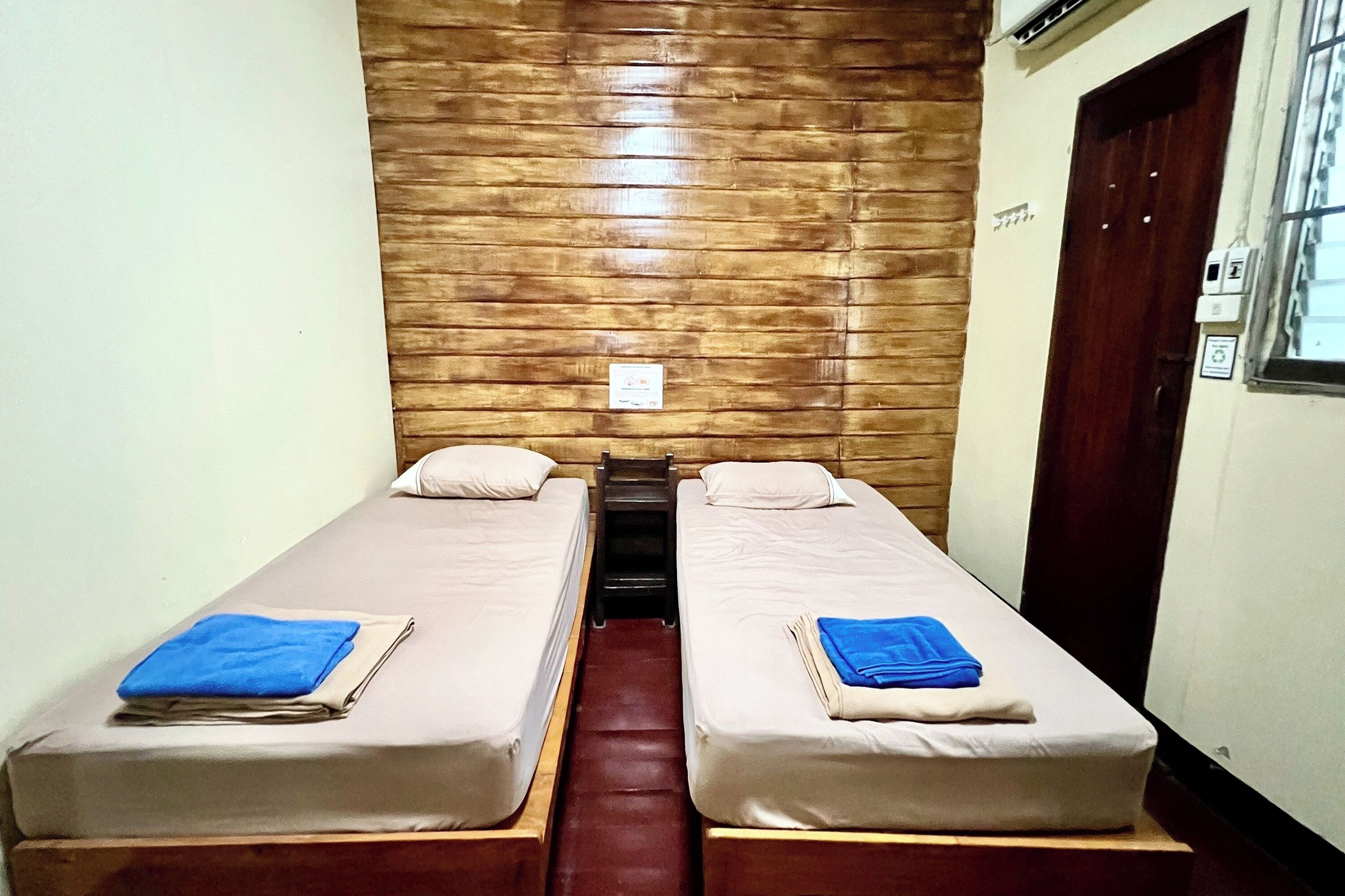 A room with two single beds, wooden paneling on the walls, a small table between the beds, and a door to the right.